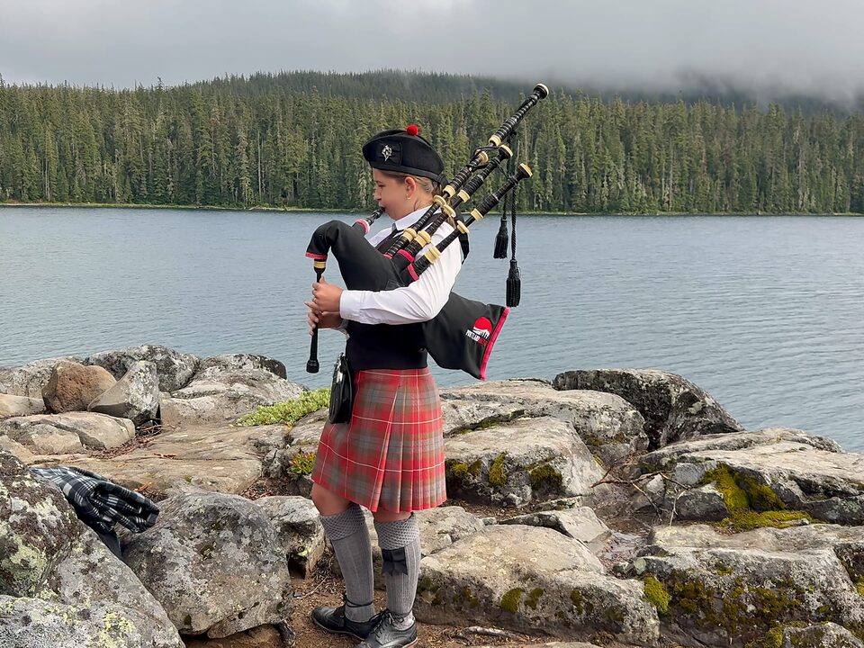 Charlotte Burslem playing bagpipes beside a mountain lake